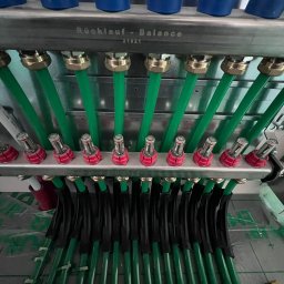 TECHNOLOGY BAU SPÓŁKA Z OGRANICZONĄ ODPOWIEDZIALNOŚCIĄ - Rozdzielacz ogrzewania podłogowego z zielonymi rurami i elementami regulacyjnymi, widoczne napisy 'Rücklauf - Balance 21821' na metalowej obudowie.