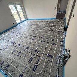 TECHNOLOGY BAU SPÓŁKA Z OGRANICZONĄ ODPOWIEDZIALNOŚCIĄ - Białe rury systemu ogrzewania podłogowego marki Schutz Pro ułożone na szarej macie izolacyjnej z niebieską taśmą brzegową w jasnym, nowym pomieszczeniu z oknami.