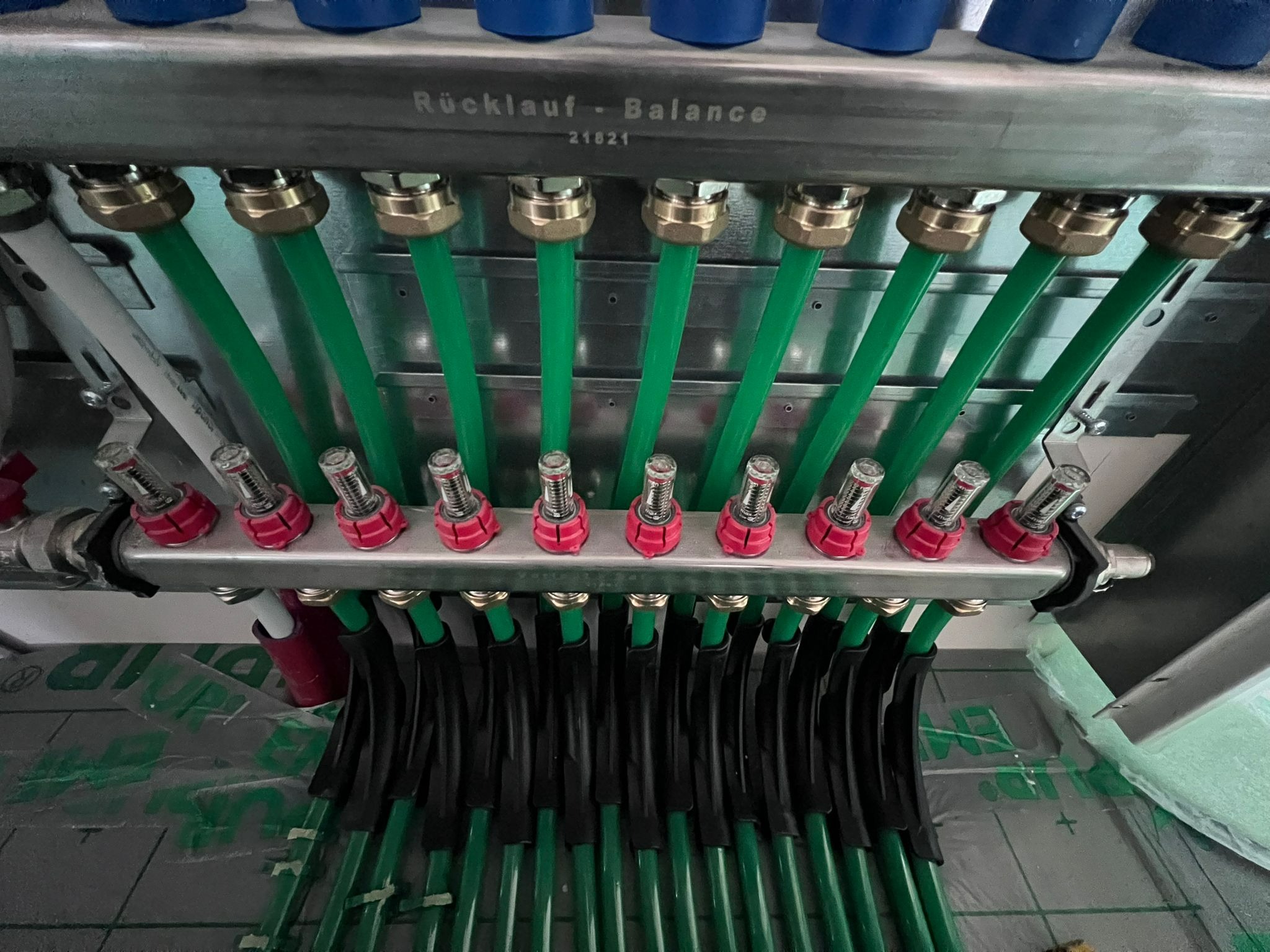 Rozdzielacz ogrzewania podłogowego z zielonymi rurami i elementami regulacyjnymi, widoczne napisy 'Rücklauf - Balance 21821' na metalowej obudowie.
