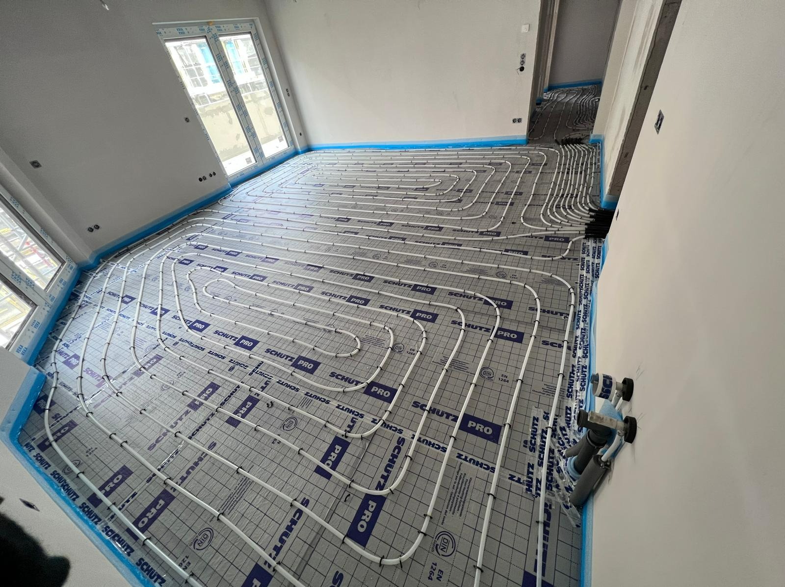 Białe rury systemu ogrzewania podłogowego marki Schutz Pro ułożone na szarej macie izolacyjnej z niebieską taśmą brzegową w jasnym, nowym pomieszczeniu z oknami.