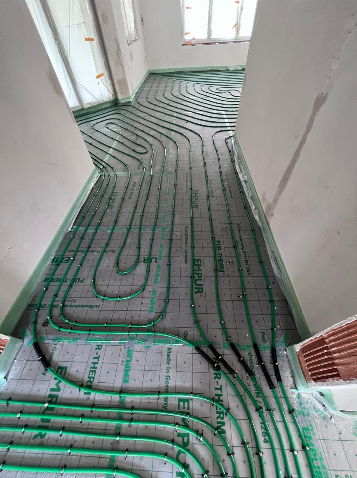 Montaż ogrzewania podłogowego: zielone rurki układane spiralnie na szarej macie izolacyjnej z widocznymi napisami 'PUR-THERM' i 'EMPUR', ściany w surowym stanie.