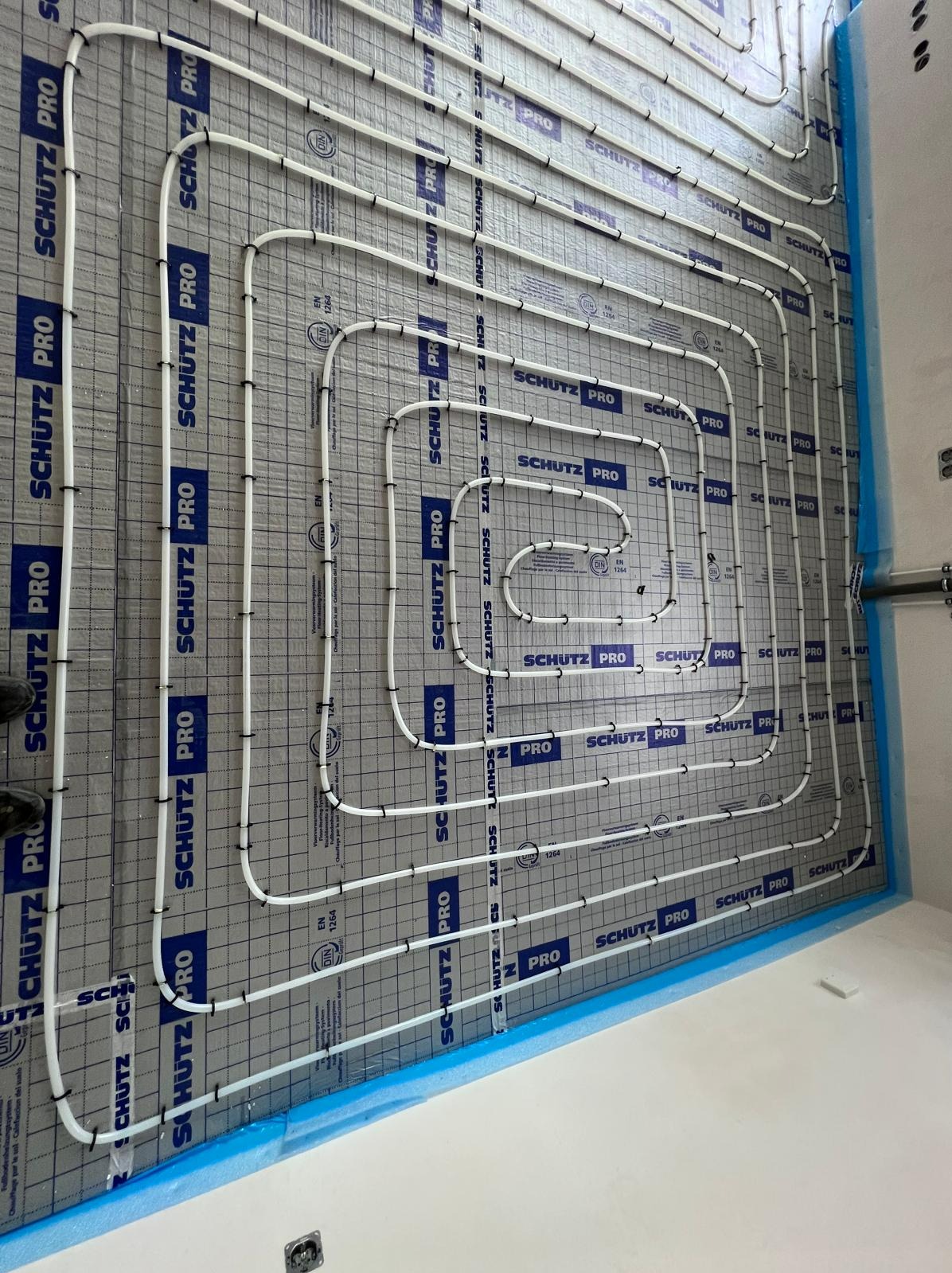 Białe rury ogrzewania podłogowego marki Schutz Pro ułożone spiralnie na szarej macie grzewczej z czarną siatką, przygotowane do zalania wylewką.