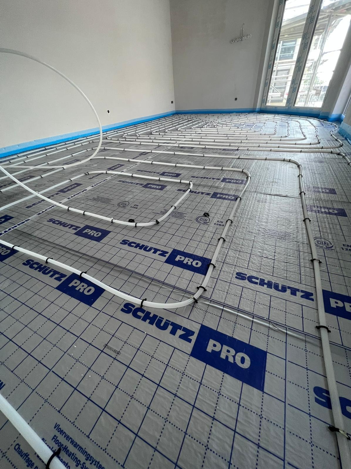 Białe rury systemu ogrzewania podłogowego marki Schutz Pro ułożone na folii z nadrukowaną siatką i logo, widoczne czarne opaski zaciskowe, świeżo wykończone wnętrze z oknem w tle.