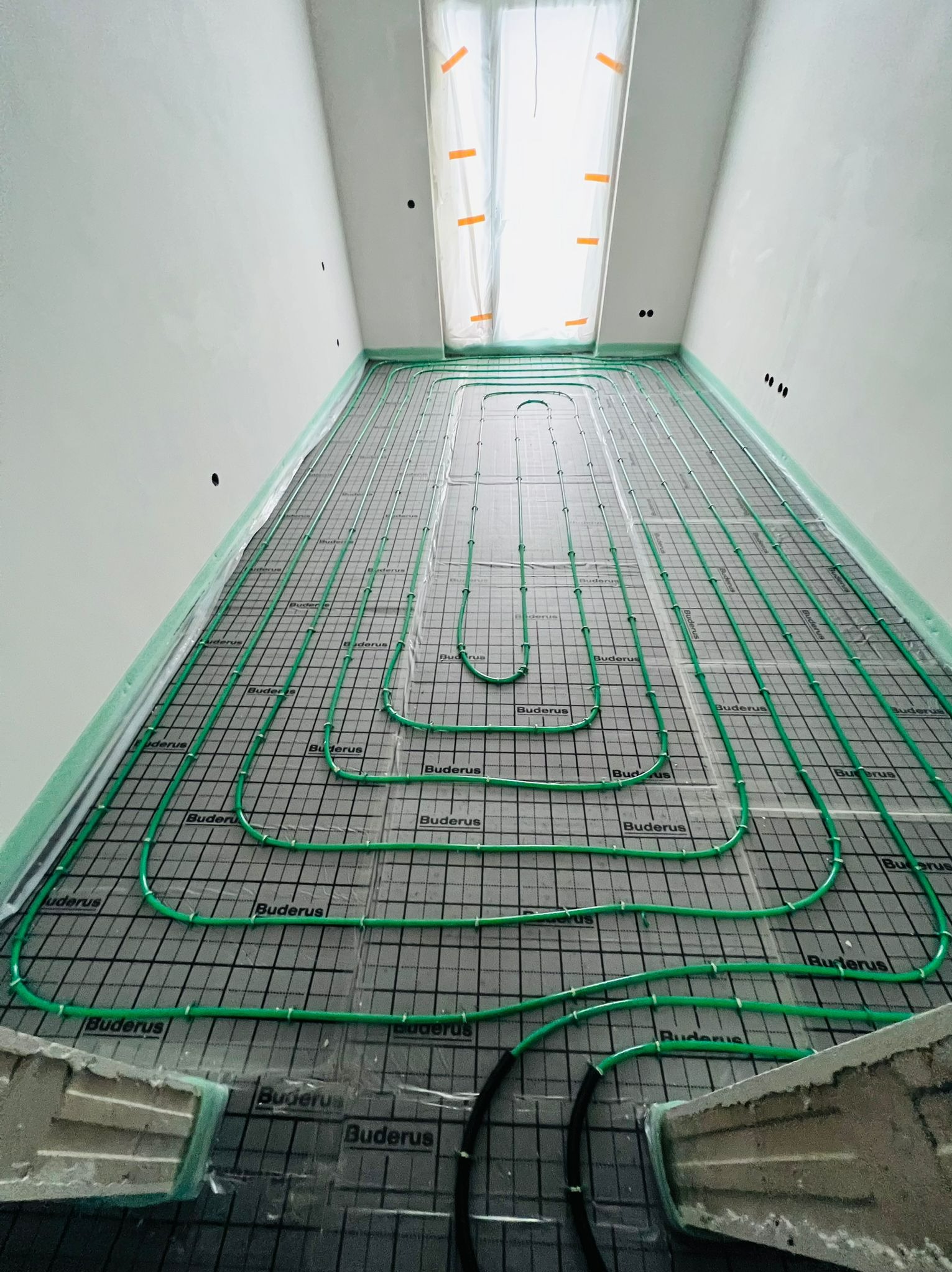 Widok z góry na rozłożone rury ogrzewania podłogowego marki Buderus na czarnej macie z siatką, zabezpieczone folią malarską przy ścianach i oknie, w surowym stanie wykończenia.