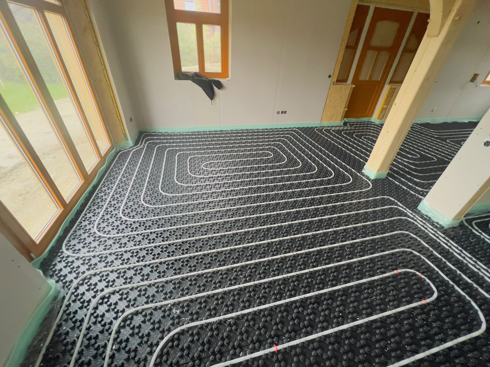 Białe rury ogrzewania podłogowego ułożone na czarnej izolacji w surowym wnętrzu z drewnianymi oknami i słupami, widok z góry.