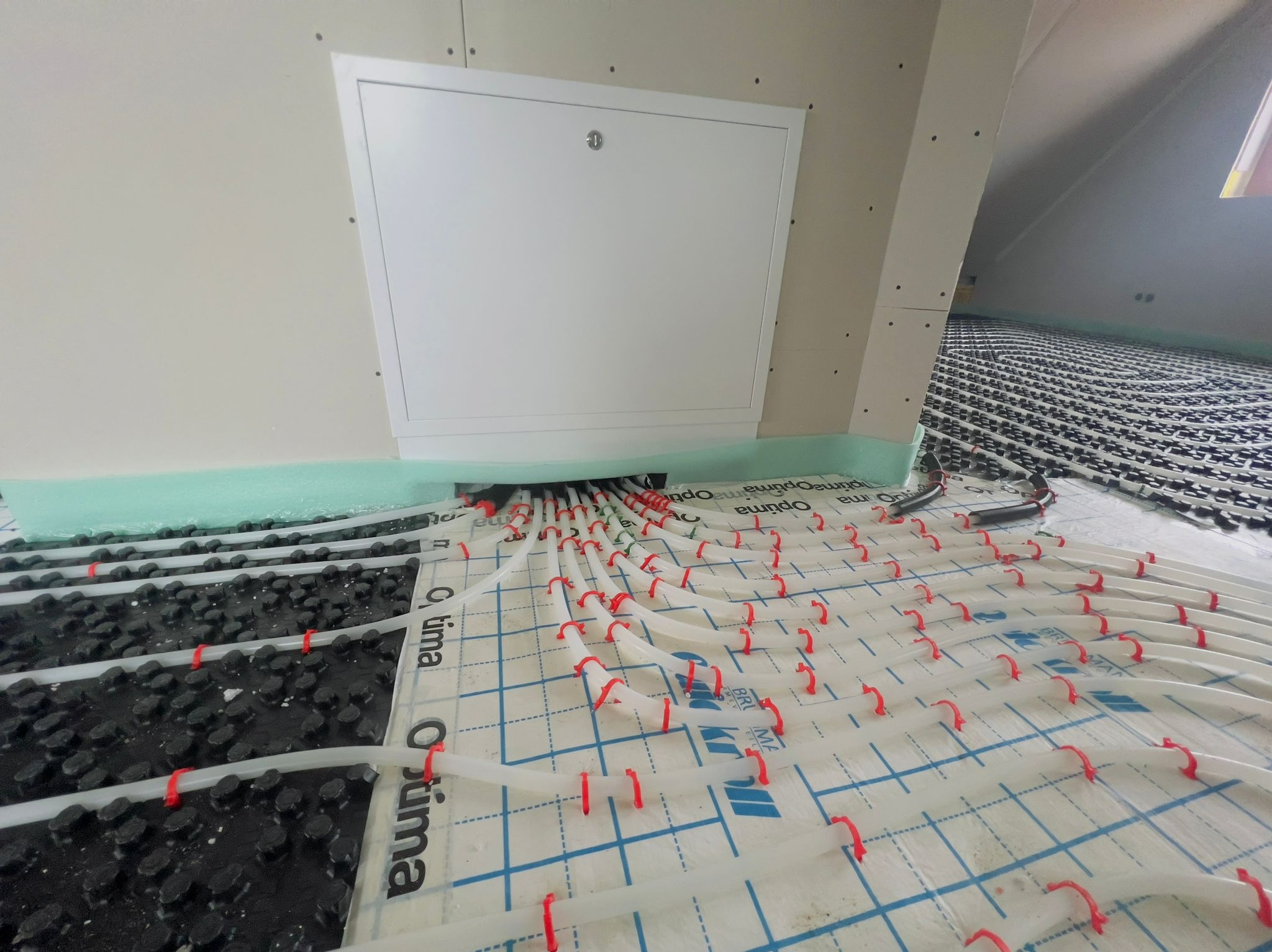 Białe rury ogrzewania podłogowego mocowane czerwonymi uchwytami na folii z nadrukiem, widoczne czarne elementy systemu, biała ściana z włazem rewizyjnym w skosie poddasza.