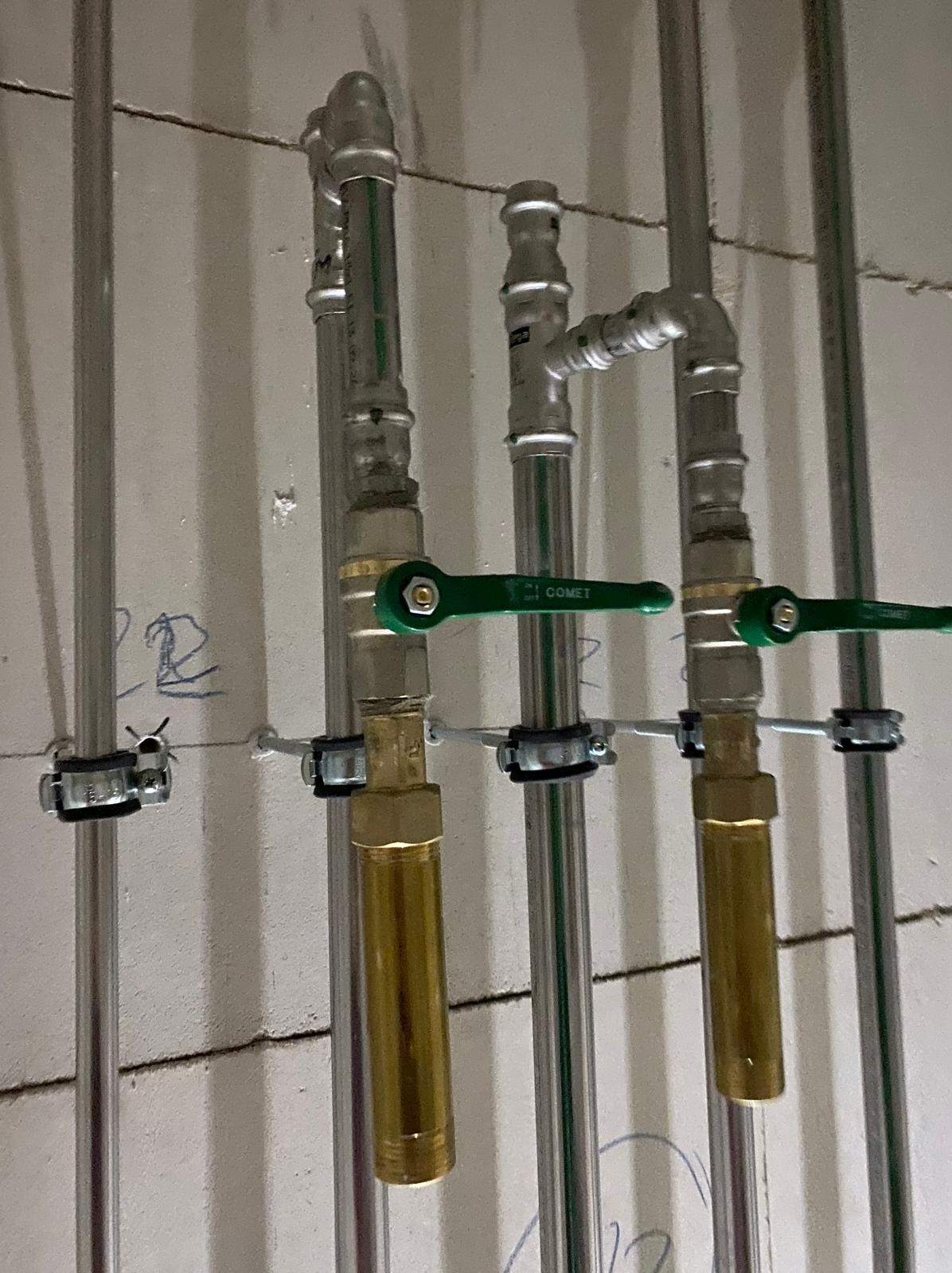 Szare rury instalacyjne z zielonymi zaworami Comet i mosiężnymi elementami, przymocowane do ściany klamrami, widoczne niebieskie napisy na ścianie.