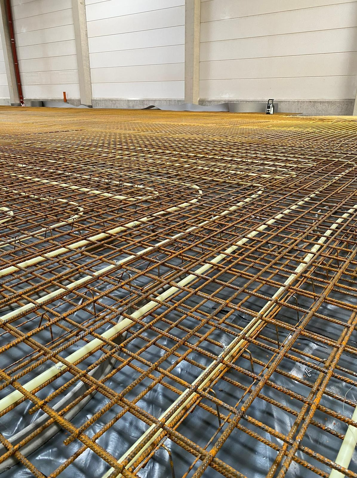 Siatka zbrojeniowa z rurami ogrzewania podłogowego na folii izolacyjnej, widok z góry w hali przemysłowej.