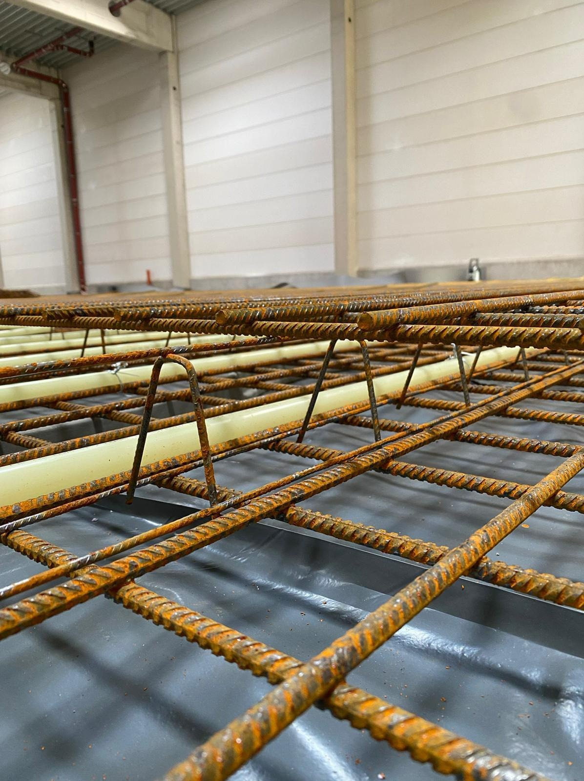 Siatka zbrojeniowa z rurami pod ogrzewanie podłogowe, ułożona na folii izolacyjnej w hali produkcyjnej, widoczne zardzewiałe pręty i białe rury.