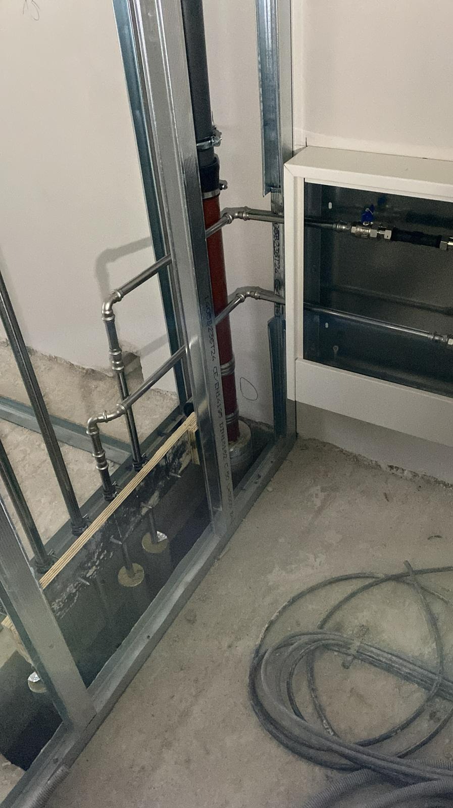 Instalacja grzewcza w trakcie montażu: rury miedziane łączące pionową rurę zasilającą z szafką rozdzielczą, widoczne elementy mocujące instalację do ściany i podłogi, surowy stan wykończenia...