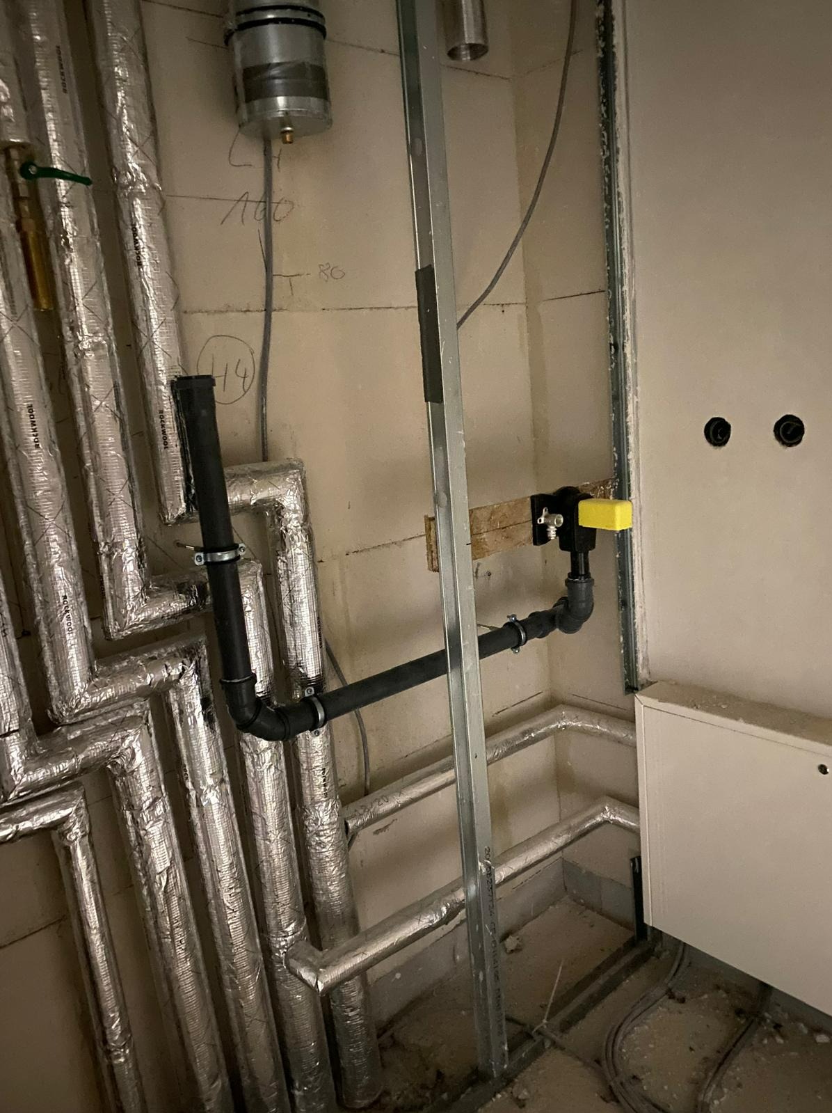 Szare rury izolowane folią aluminiową oraz czarne rury z zaworem odcinającym z żółtą nakładką, montowane w surowym pomieszczeniu z metalowym profilem i instalacją elektryczną w ścianie.