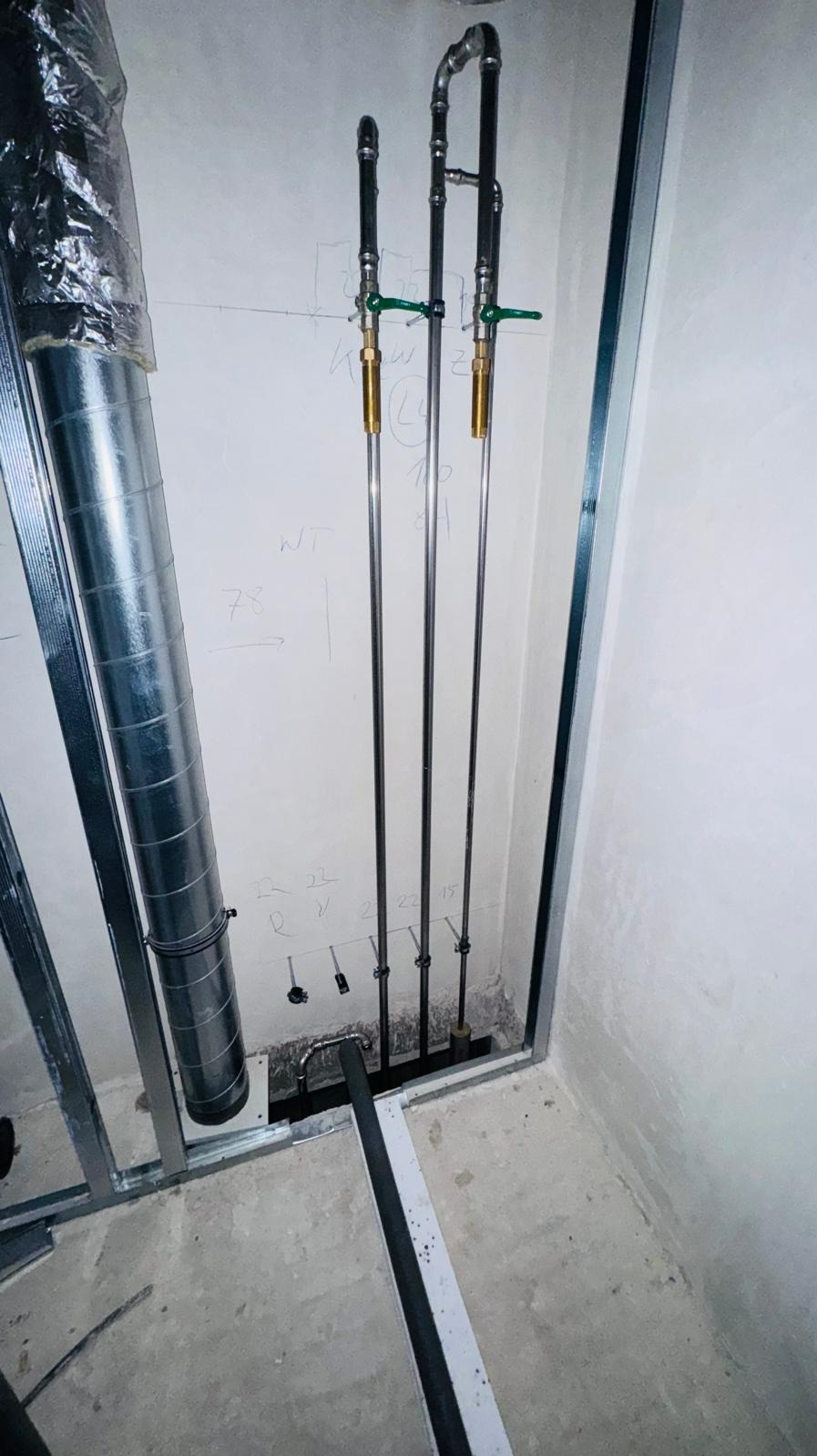 Surowe wnętrze w trakcie prac wykończeniowych: pionowe rury instalacji wodnej z zaworami odcinającymi, widoczne elementy konstrukcji ścian działowych oraz fragment kanału wentylacyjnego.