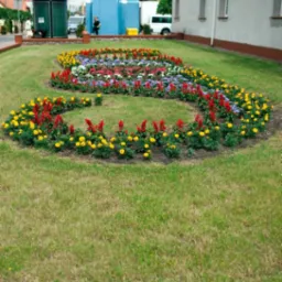 Ogród miejski z kolorowymi kwiatami ułożonymi w kształt litery S, na tle budynku i toalety publicznej.