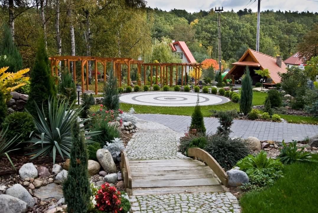 Kompleksowo zaprojektowany ogród z drewnianym mostkiem nad strumieniem, prowadzącym do okrągłego placu z pergolą, otoczony różnorodną roślinnością i elementami małej architektury.