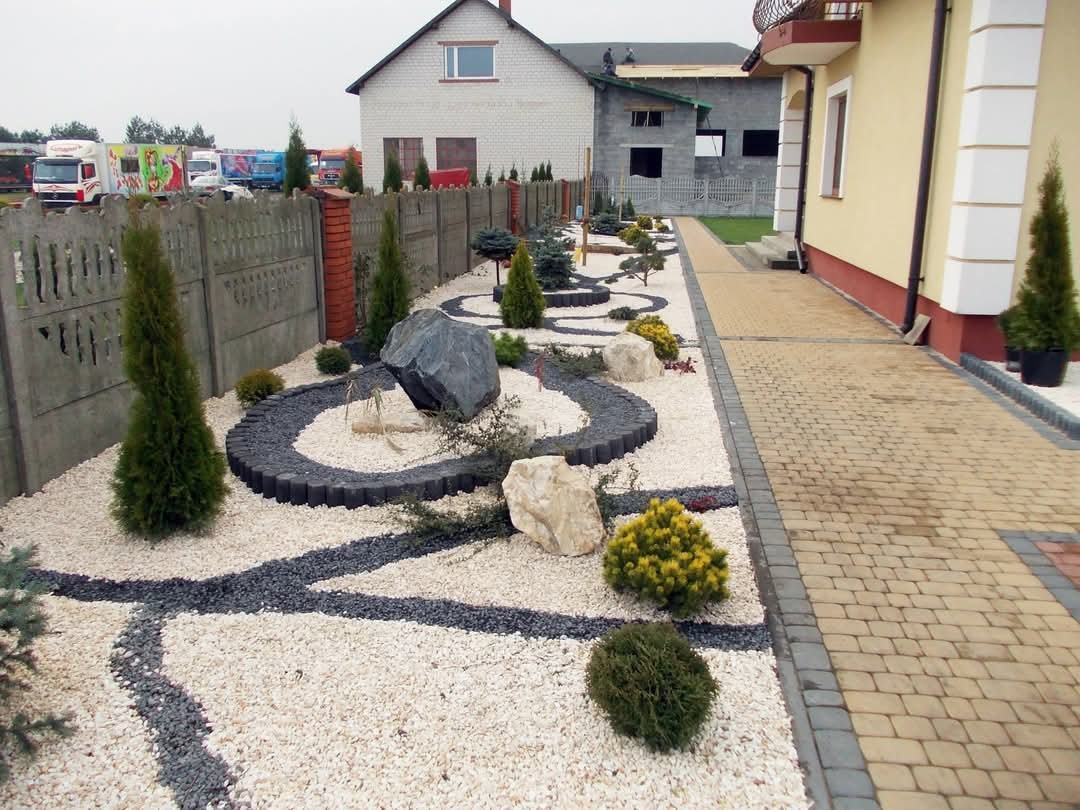 Ogród z białym żwirem, ścieżkami z czarnego grysu i dużymi kamieniami, obok brukowany chodnik i dom, w tle budynek w trakcie budowy.