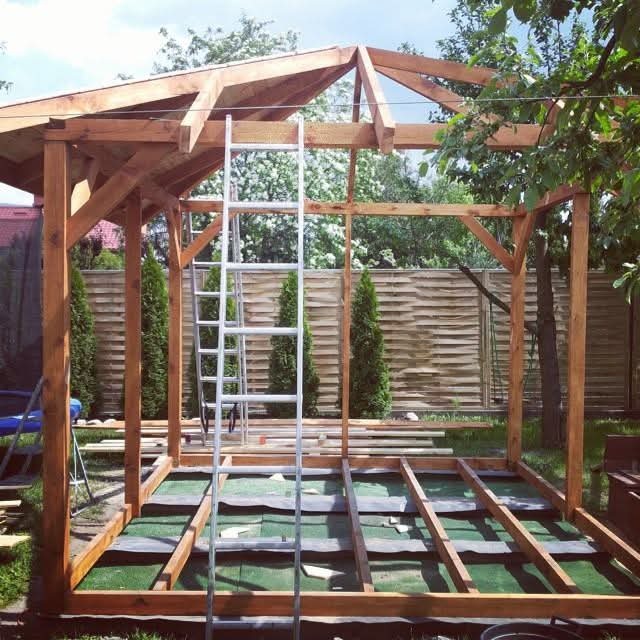 Drewniana konstrukcja altany ogrodowej w trakcie budowy, widoczny szkielet dachu i podłogi, drabina oparta o konstrukcję, w tle tuje i płot.