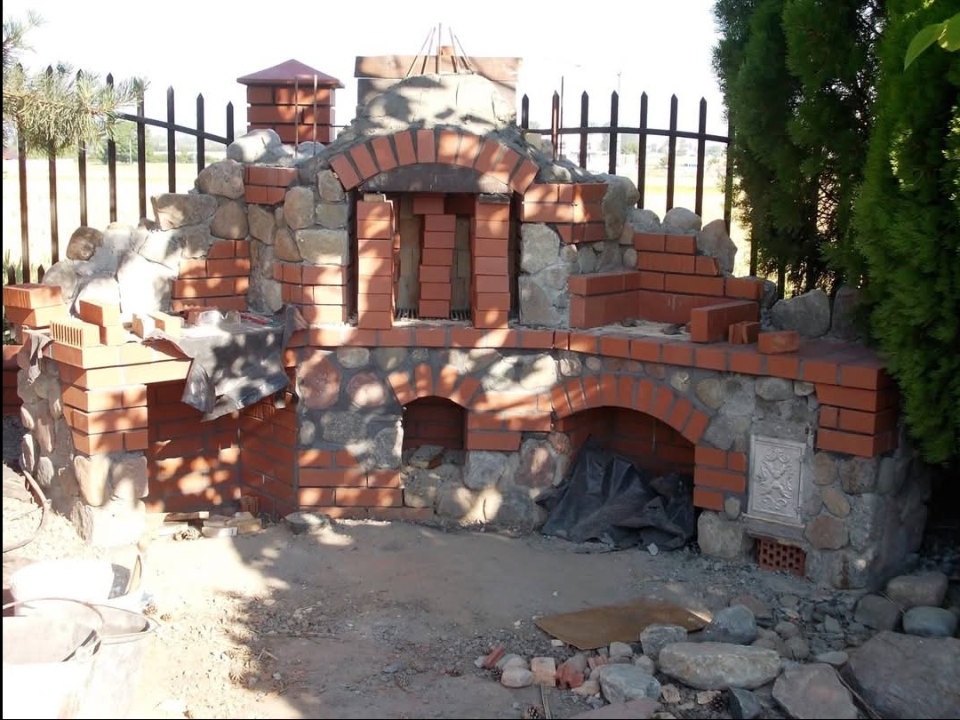 Budowa murowanego grilla ogrodowego z cegły i kamienia, widoczne elementy konstrukcyjne w trakcie wznoszenia, teren wokół jeszcze niezagospodarowany.