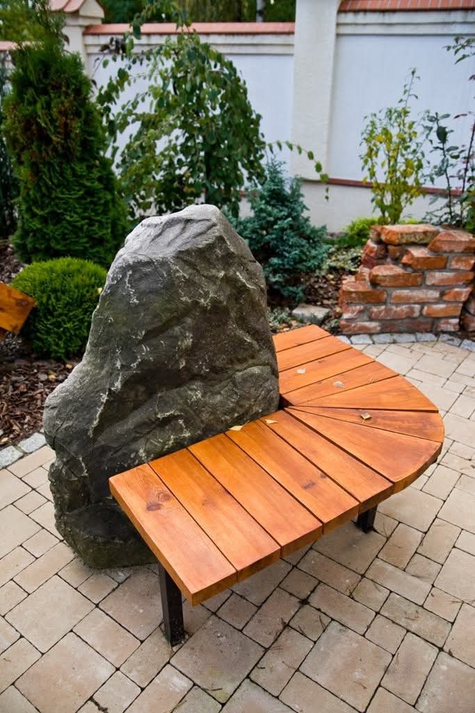 Kamienna ławka ogrodowa z drewnianym siedziskiem, otoczona zielenią i brukowaną ścieżką, na tle białego muru.