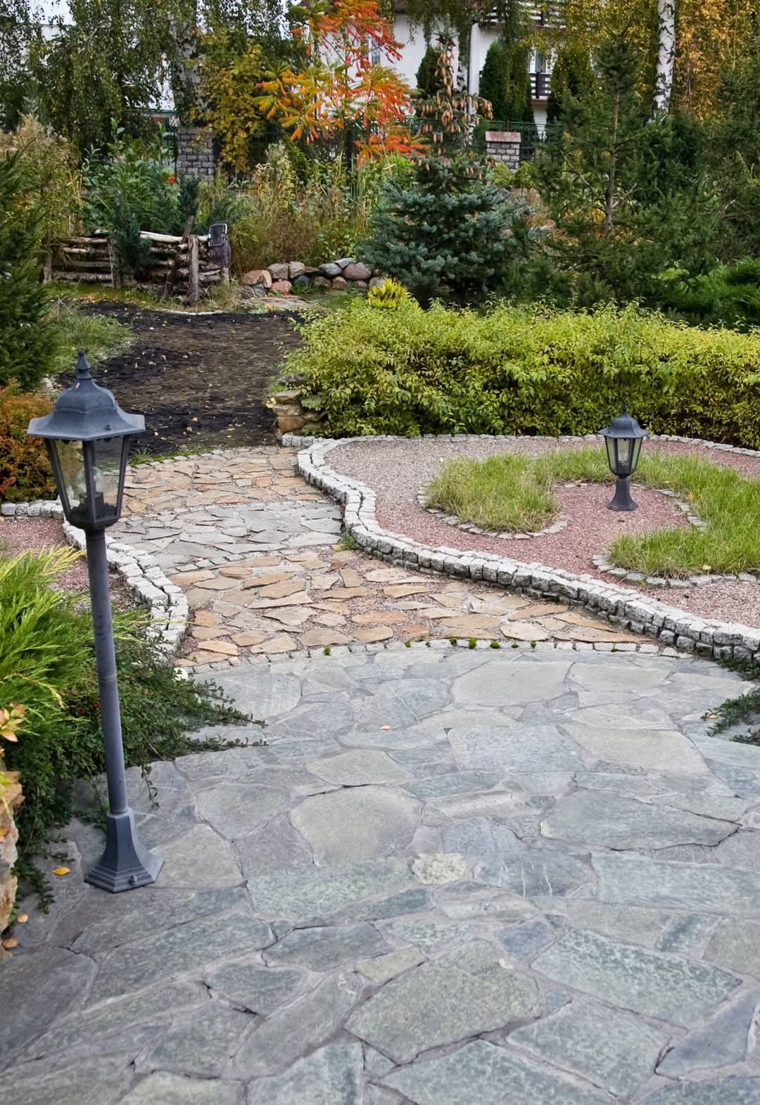 Ogród z kamienną ścieżką z nieregularnych płyt, oświetlony dwoma czarnymi latarniami, z elementami rabat obsypanych czerwonym żwirem i niskimi krzewami, w tle drzewa.