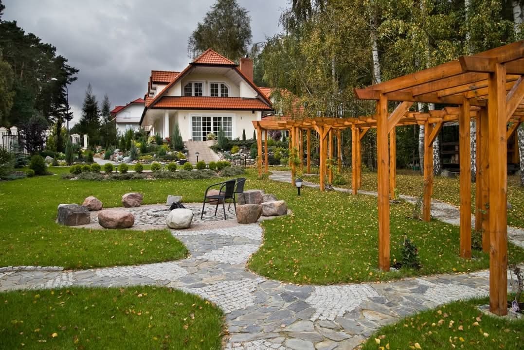 Przestronny ogród z pergolą, ścieżką z kamienia naturalnego prowadzącą do paleniska otoczonego skałami, na tle białego domu z czerwonym dachem.