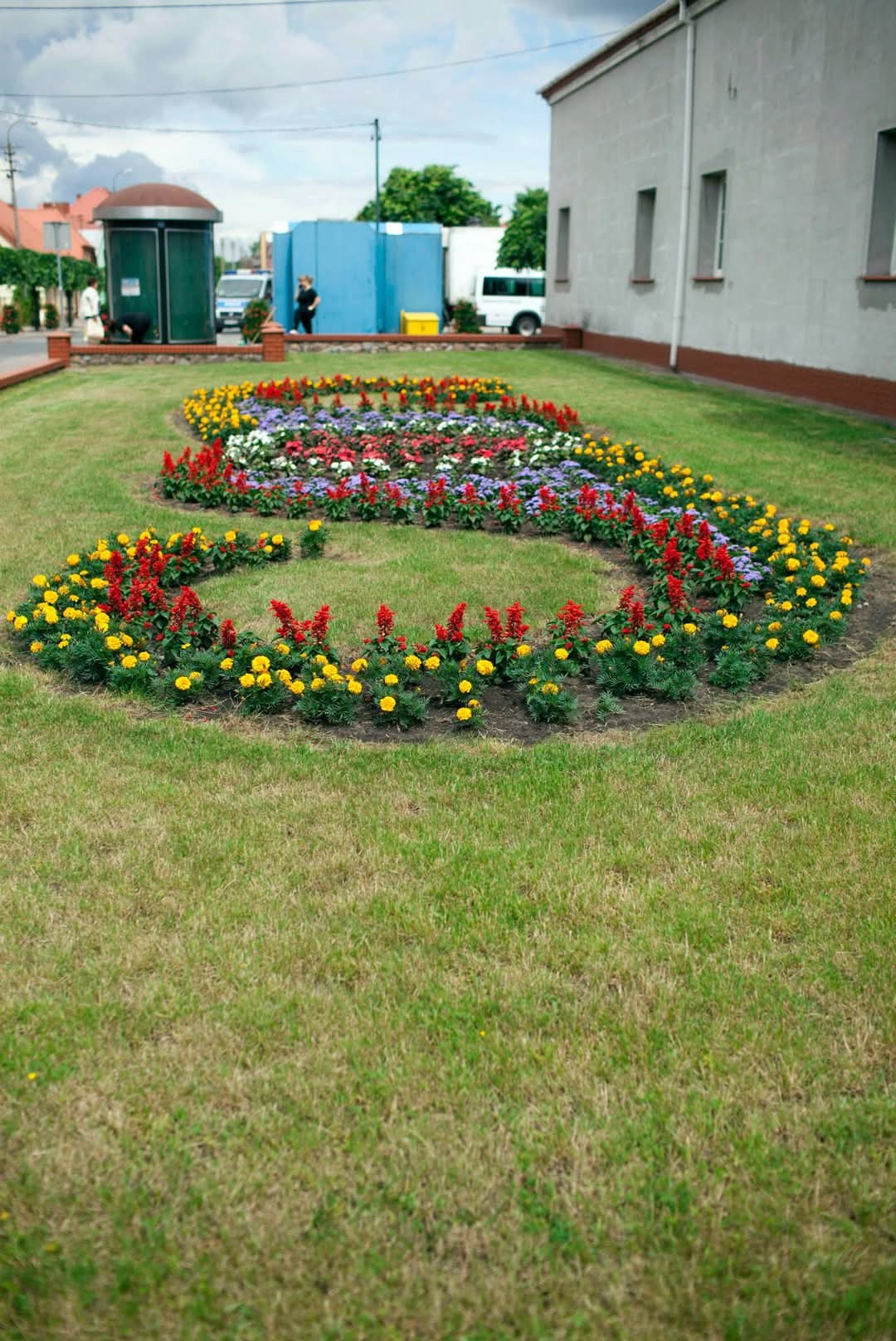 Ogród miejski z kolorowymi kwiatami ułożonymi w kształt litery S, na tle budynku i toalety publicznej.