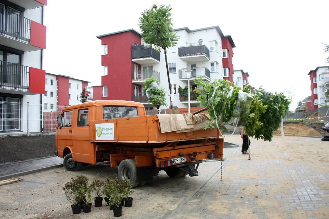 Pomarańczowy samochód dostawczy z przyczepą pełną drzewek i roślin, przygotowanych do zasadzenia na terenie nowego osiedla mieszkaniowego.