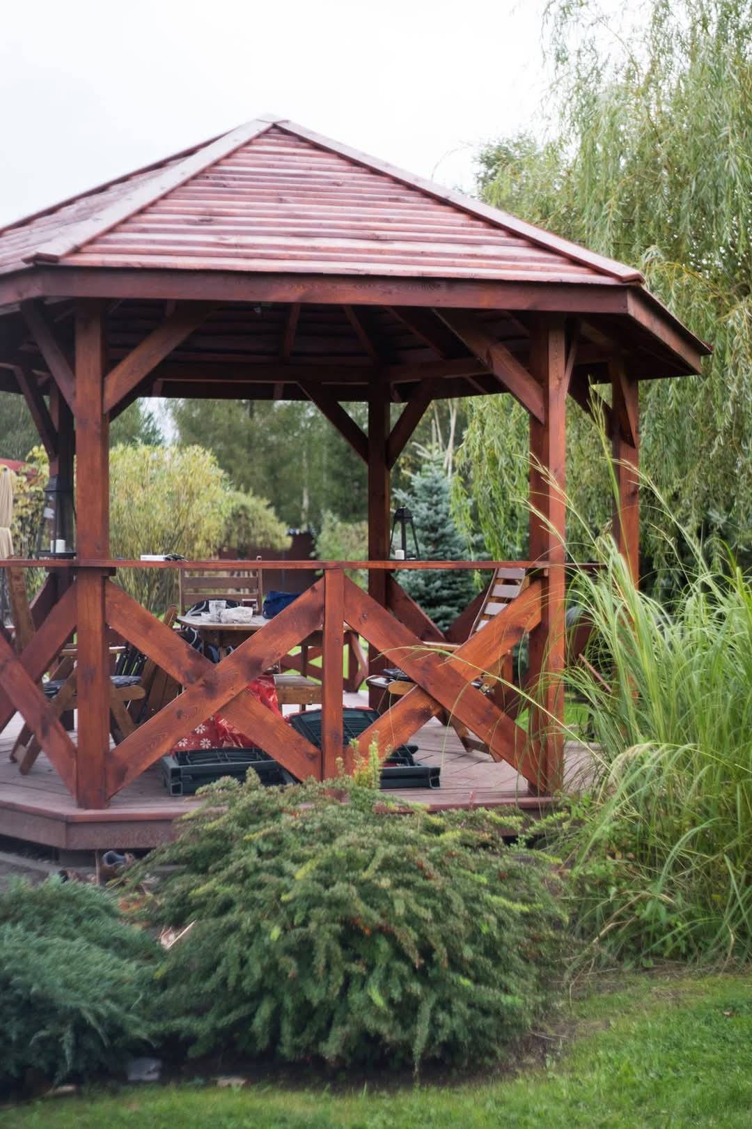 Drewniana altana ogrodowa z brązowym dachem, otoczona zielenią, w tle drzewa i krzewy.