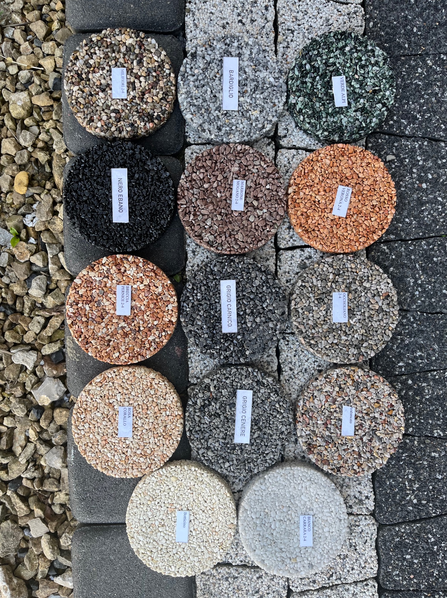 Prezentacja różnych wariantów kolorystycznych i faktur kruszywa żywicznego na okrągłych próbkach z etykietami, ułożonych na szarej kostce brukowej i żwirze.