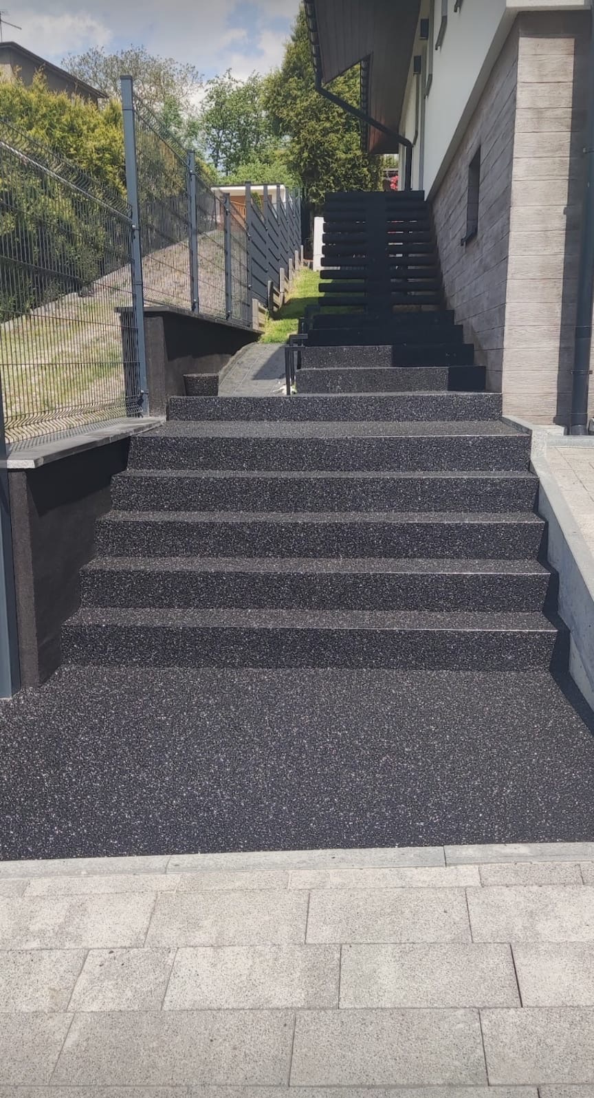 Zewnętrzne schody pokryte czarną, drobną posadzką żywiczną z widoczną fakturą, prowadzące do domu z elewacją imitującą drewno.