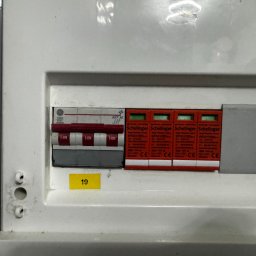 Oskar Kasprowicz - Rozdzielnica elektryczna z wyłącznikami i zabezpieczeniami przeciwprzepięciowymi marki Schelinger, widoczne oznaczenia i parametry techniczne. Żółta naklejka z numerem '19'.