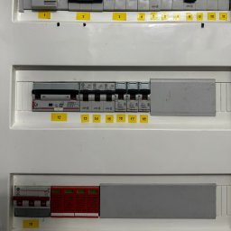 Oskar Kasprowicz - Rozdzielnica elektryczna z bezpiecznikami i oznaczeniami, widok z bliska. Elementy instalacji elektrycznej wewnątrz skrzynki, z numeracją na żółtych naklejkach.