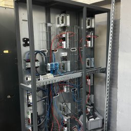 Kastal Electric - Szafa sterownicza w trakcie montażu, widoczne liczne przewody, moduły i elementy mocujące. Szara obudowa z metalową ramą, częściowo okablowana, na tle szarej ściany.