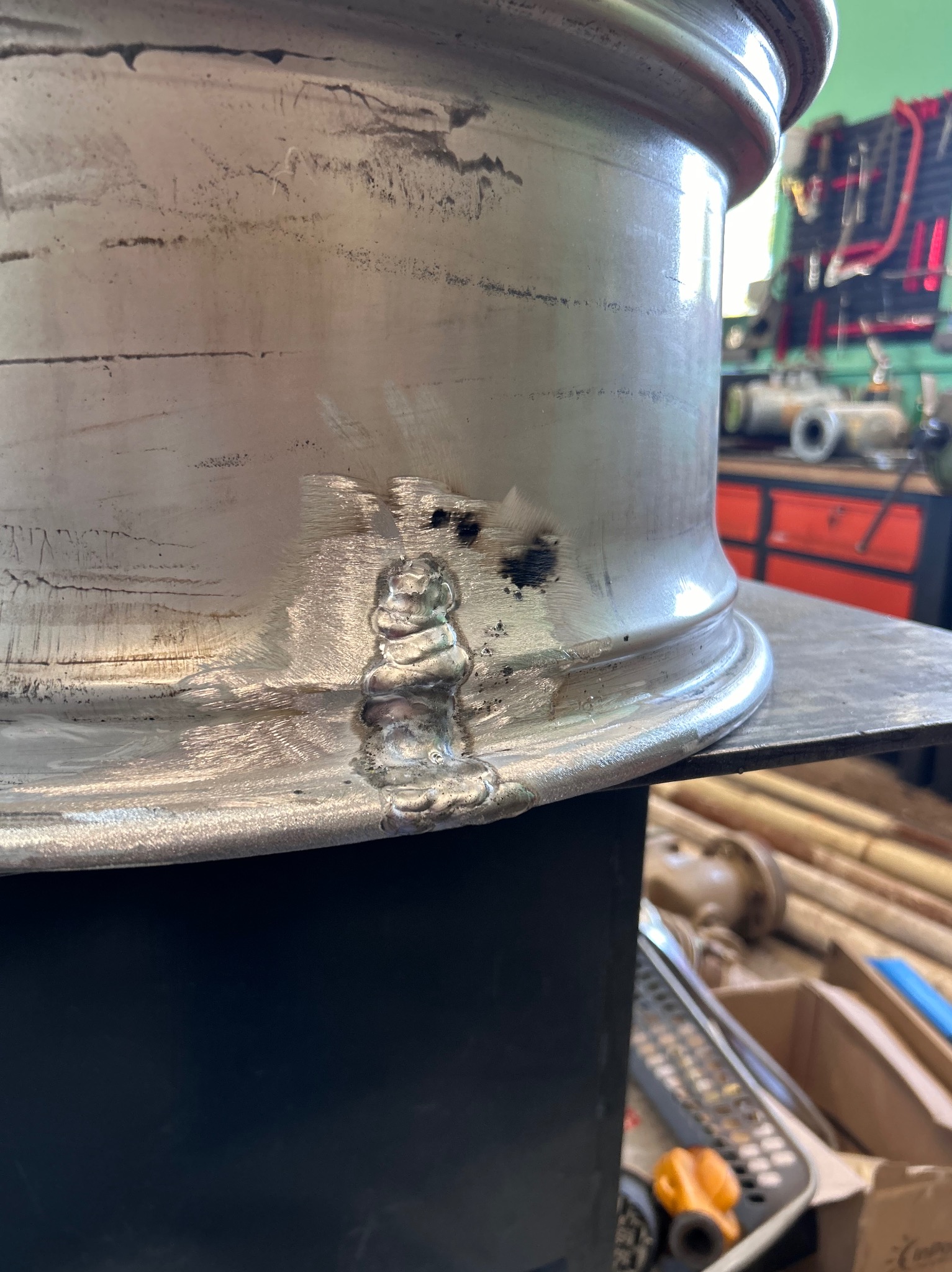 Fragment felgi aluminiowej po spawaniu, widoczne ślady po spawarce i niedoskonałości spoiny, warsztat w tle.