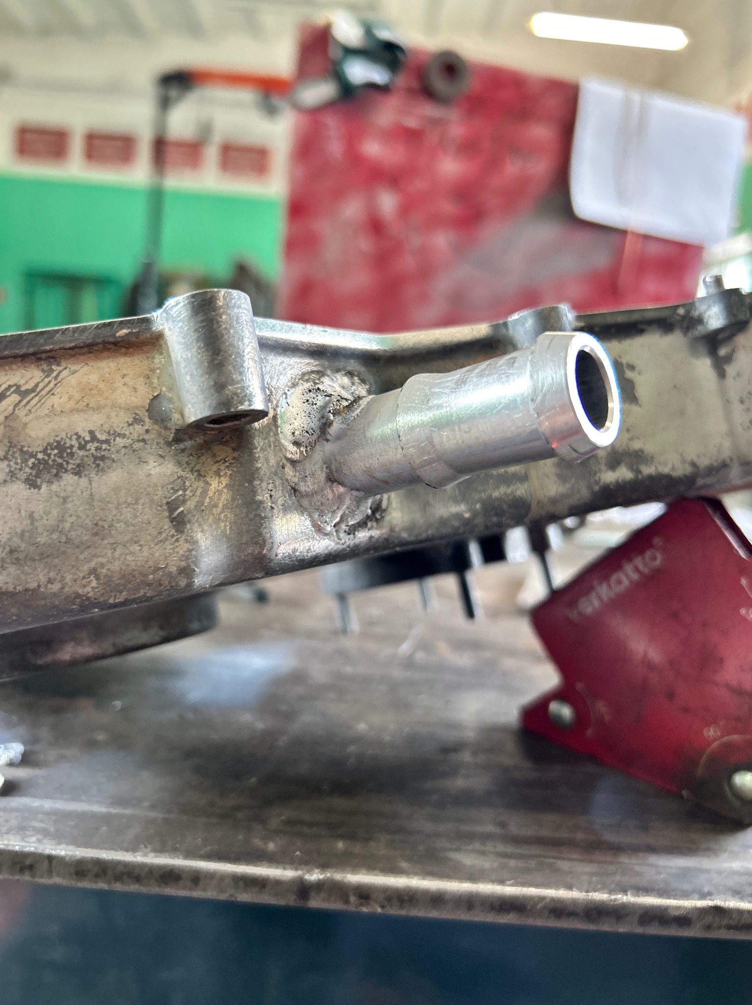 Szczegółowe ujęcie elementu metalowego z przyspawaną rurką, widoczne ślady spawania i obróbki, umieszczony na metalowym stole warsztatowym z czerwonym imadłem w tle.