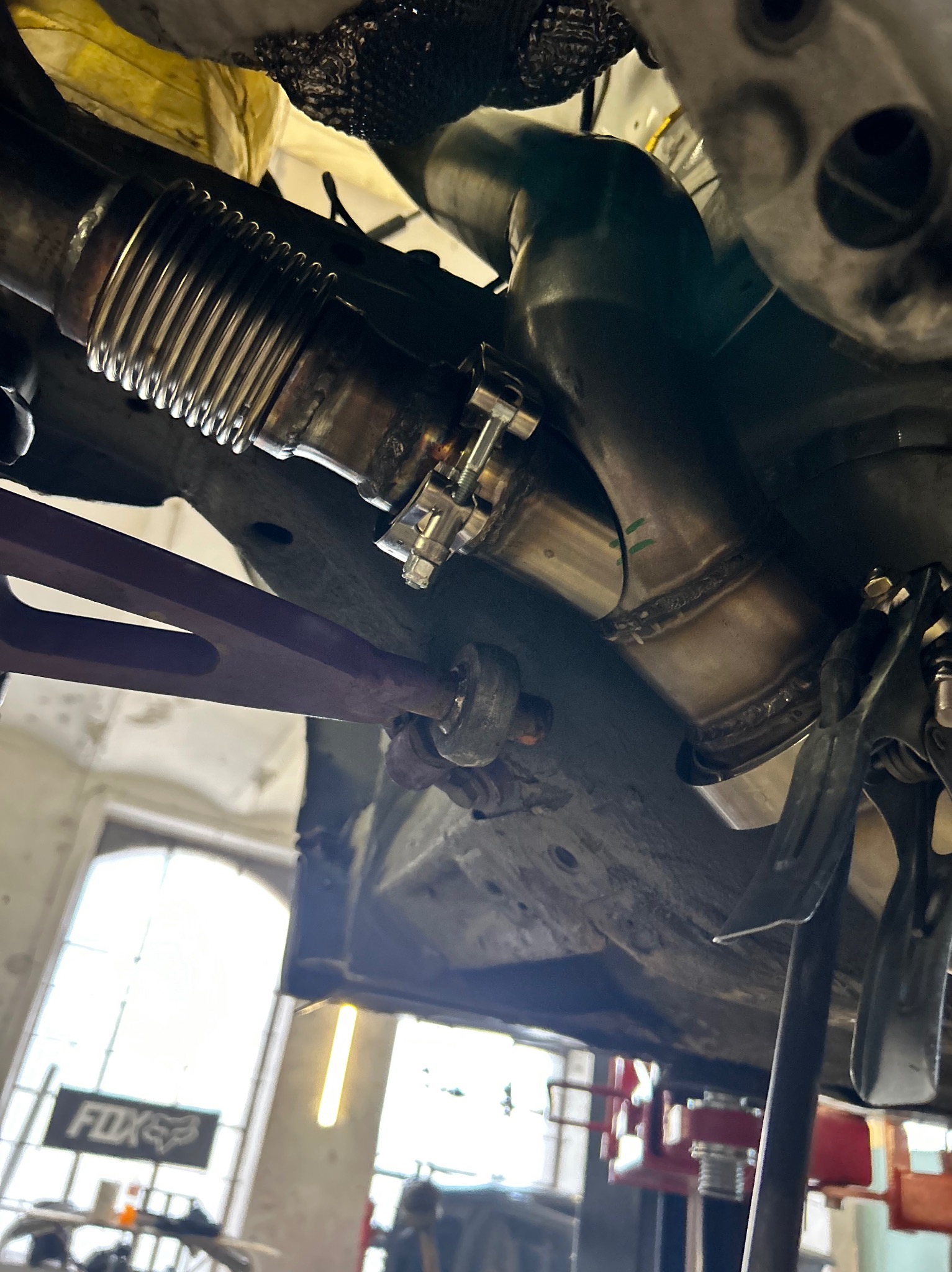 Spawany układ wydechowy samochodu od dołu, z widoczną elastyczną rurą, spawami i elementami mocującymi, w warsztacie samochodowym z podnośnikiem.