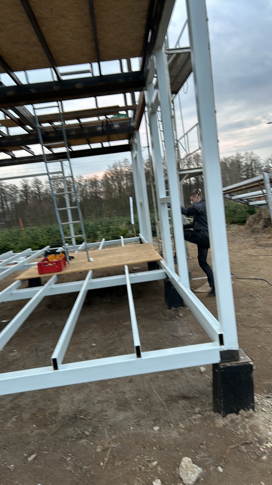 Montaż szkieletu metalowej konstrukcji, widoczny pracownik w masce wspinający się po białych profilach, narzędzia w czerwonej skrzynce, surowy krajobraz w tle.