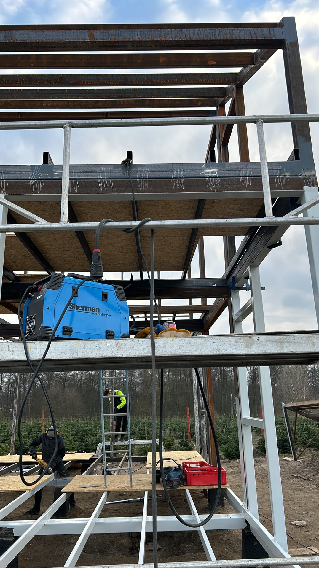 Konstrukcja stalowa w trakcie spawania, widoczny spawacz w czarnym ubraniu i niebieska spawarka Sherman na podwyższeniu, drugi pracownik na drabinie w tle, elementy konstrukcji z surowej stali...