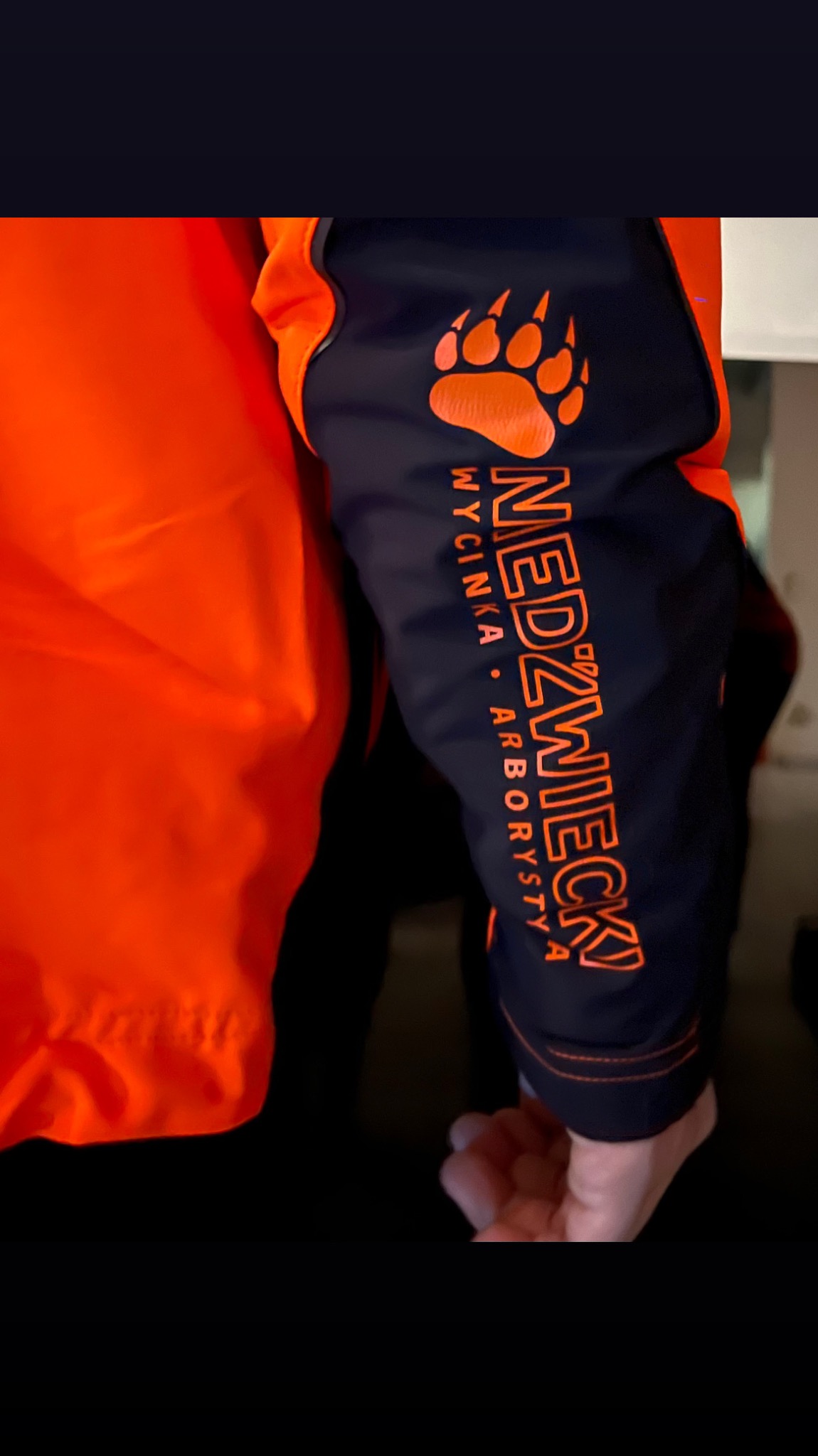 Rękaw roboczy w kolorze granatowym z pomarańczowym logo niedźwiedziej łapy i napisem 'NEDZWIECKY WYCINKA ARBORYSTA', część pomarańczowej kurtki roboczej.