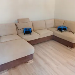 Narożnik po praniu tapicerki, na siedziskach ustawione wentylatory wspomagające suszenie. Widok na jasny, dwukolorowy mebel i drewnianą podłogę.