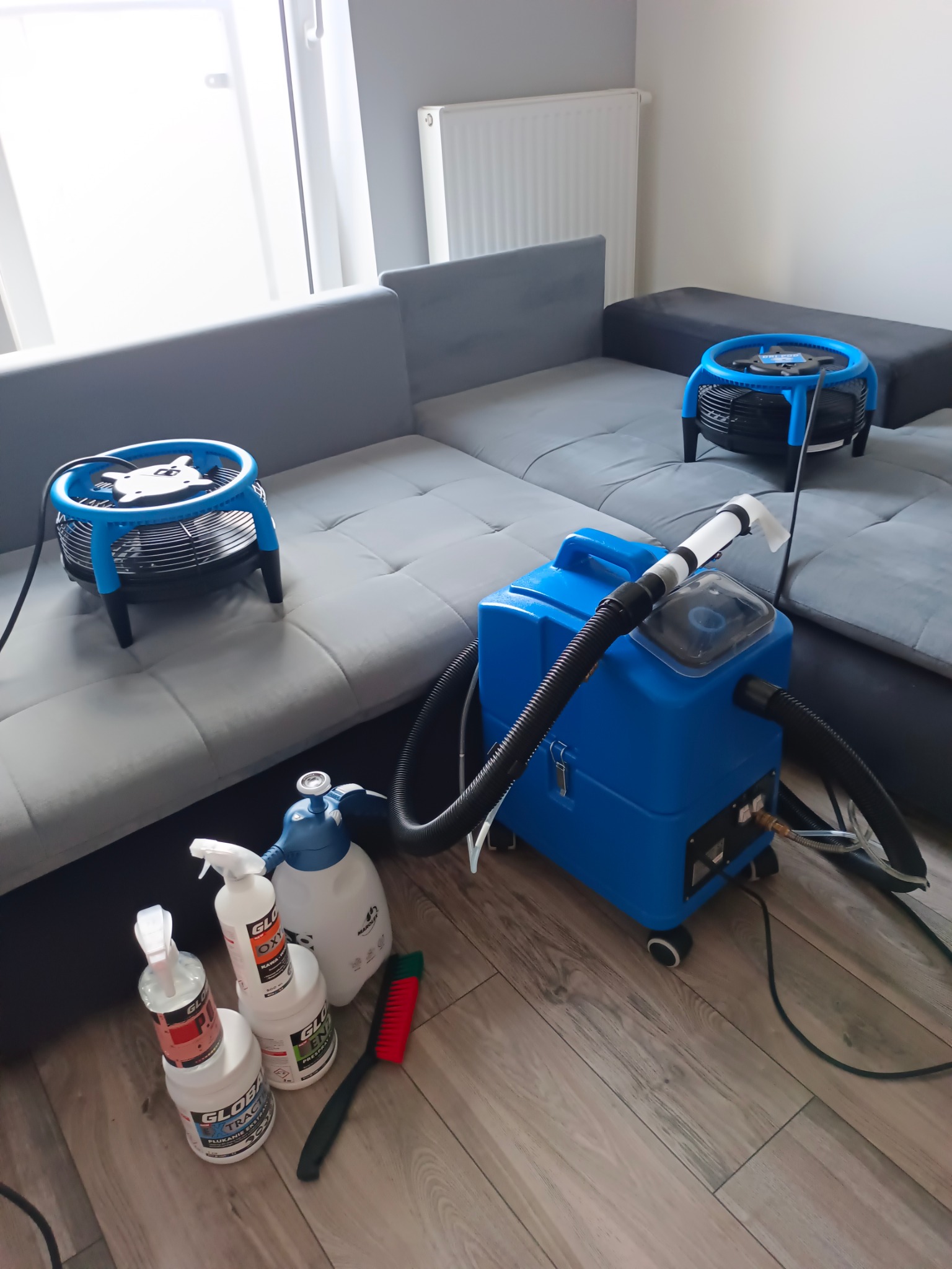 Profesjonalny sprzęt do prania tapicerki: ekstraktor, wentylatory, chemia. Widok na szarą sofę i drewnianą podłogę. Usługi czyszczenia.