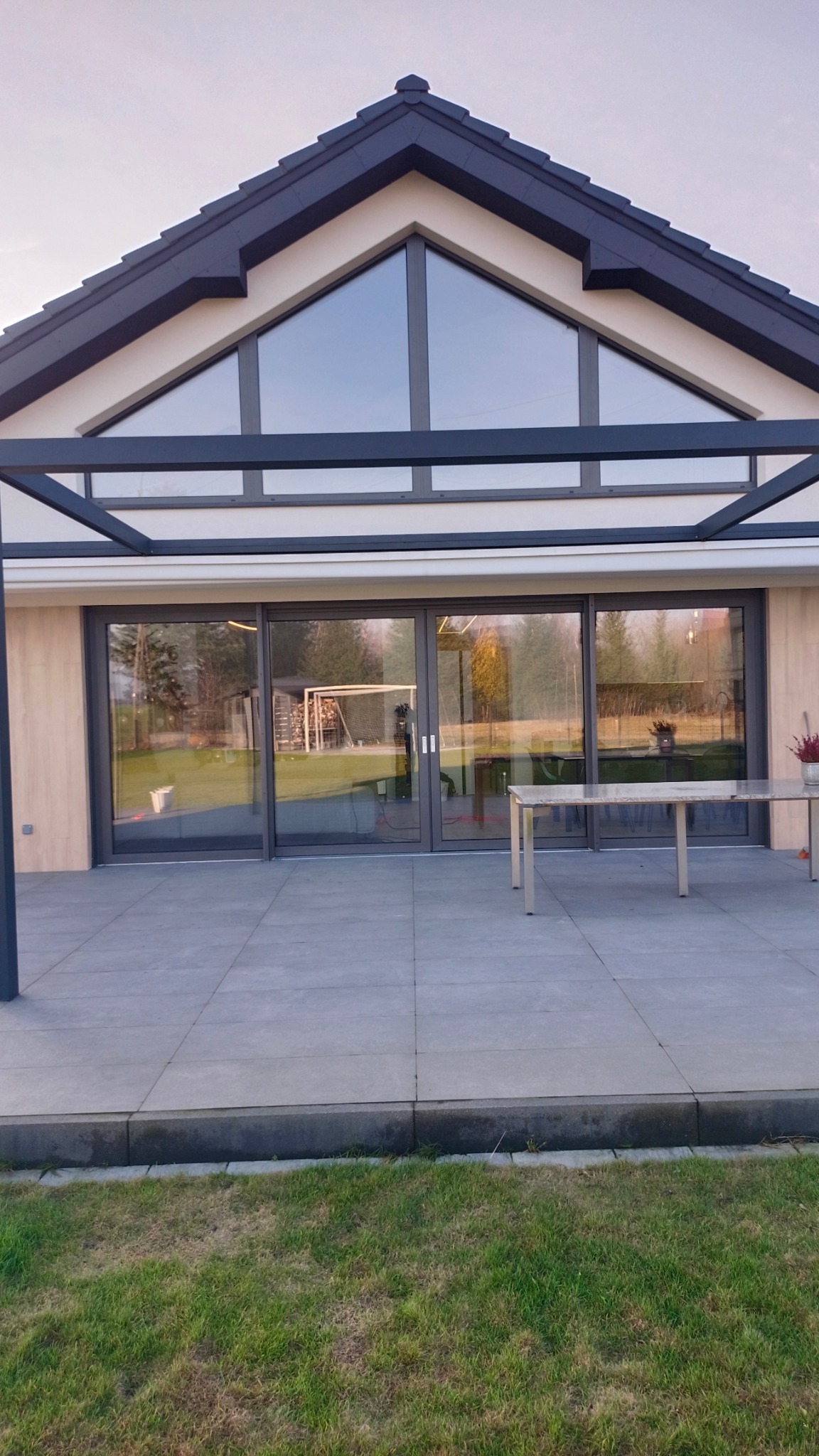 Nowoczesny dom z dużymi, przesuwnymi oknami tarasowymi i szarą kostką brukową na patio. Widok na zadbany ogród z siatką do gry w piłkę nożną.