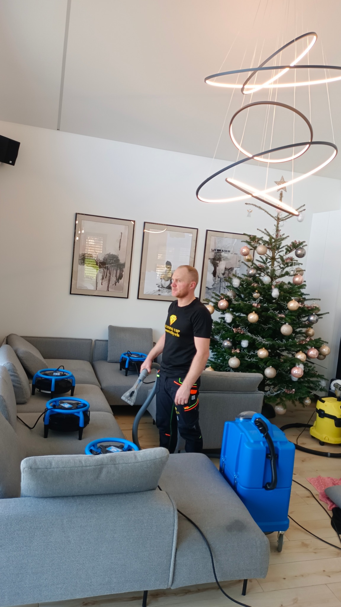 Pracownik w trakcie prania narożnika w salonie z choinką, używając profesjonalnego sprzętu. Widoczne wentylatory wspomagające suszenie tapicerki.