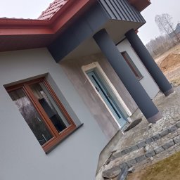 TYBER-DACH - Nowoczesny dom jednorodzinny z szarą elewacją i czerwoną dachówką, widoczne okna z drewnianymi ramami i grafitowe kolumny przy wejściu, teren wokół domu w trakcie prac wykończeniowych.