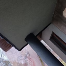 TYBER-DACH - Widok z dołu na fragment elewacji budynku w trakcie prac wykończeniowych: świeżo pomalowana kolumna wsporcza, fragment podbitki dachowej, ściana z oknem po nałożeniu warstwy tynku, widoczne ślady...