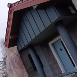 Elewacja domu w trakcie malowania, widoczne rusztowania i zabezpieczenia, nowa blaszana obudowa lukarny w kolorze grafitowym, czerwona dachówka i błękitne drzwi wejściowe.