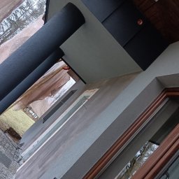 TYBER-DACH - Fragment elewacji budynku z oknem w drewnianej ramie, widoczna struktura tynku, czarne panele na dachu i rura spustowa, ujęcie z dołu w trakcie prac wykończeniowych.