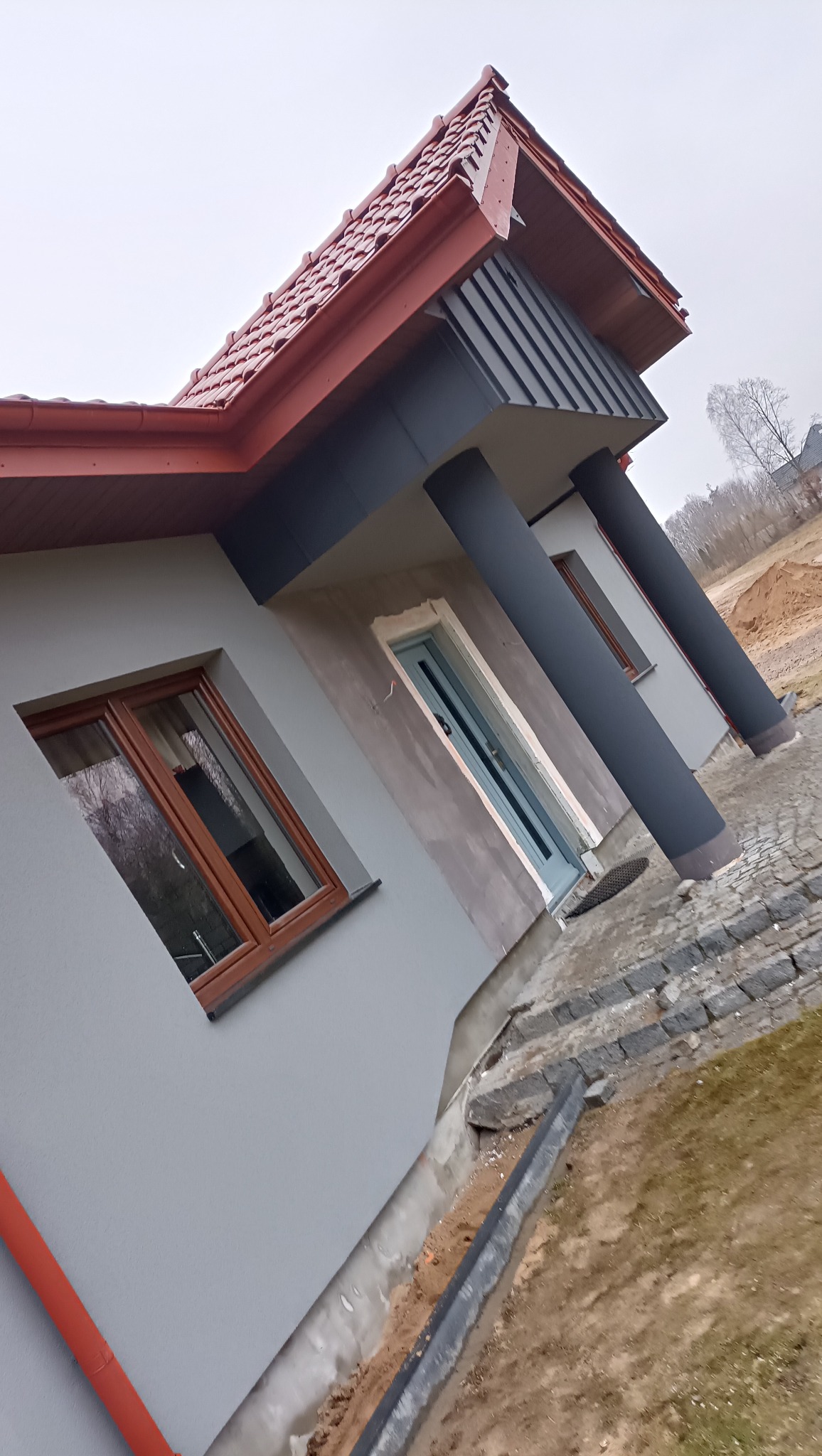 Nowoczesny dom jednorodzinny z szarą elewacją i czerwoną dachówką, widoczne okna z drewnianymi ramami i grafitowe kolumny przy wejściu, teren wokół domu w trakcie prac wykończeniowych.