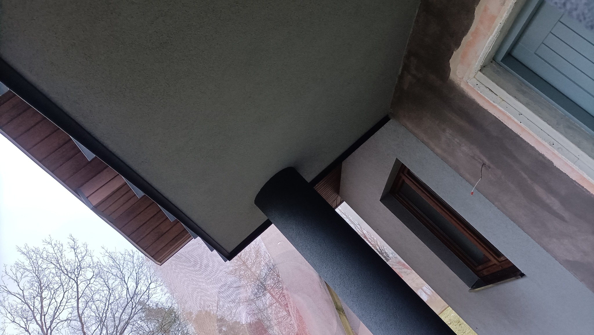 Widok z dołu na fragment elewacji budynku w trakcie prac wykończeniowych: świeżo pomalowana kolumna wsporcza, fragment podbitki dachowej, ściana z oknem po nałożeniu warstwy tynku, widoczne ślady...