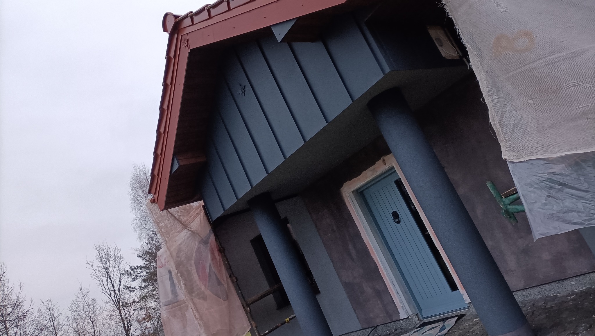 Elewacja domu w trakcie malowania, widoczne rusztowania i zabezpieczenia, nowa blaszana obudowa lukarny w kolorze grafitowym, czerwona dachówka i błękitne drzwi wejściowe.