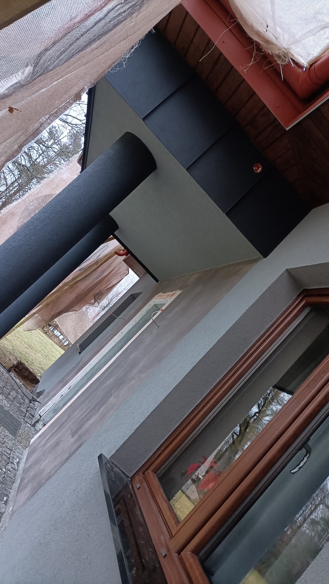 Fragment elewacji budynku z oknem w drewnianej ramie, widoczna struktura tynku, czarne panele na dachu i rura spustowa, ujęcie z dołu w trakcie prac wykończeniowych.